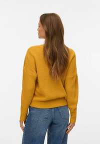 Dzeltens adīts džemperis ar vaļīgu piegriezumu, raksturīgs ar teksturētu virsmu un ribveida apkaklēm, kombinēts ar augstvēderu džinsa biksēm.