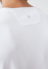 Λευκή, υφασμάτινη αθλητική μπλούζα με υφή; διαθέτει στρογγυλή λαιμόκοψη και μια ετικέτα με μινιμαλιστικό λογότυπο στην πλάτη.