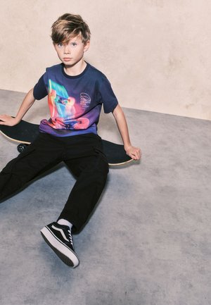 Mladý chlapec s modrýma očima sedí na betonové podlaze a drží skateboard, má na sobě barevné tričko s potiskem, černé kalhoty a tenisky.