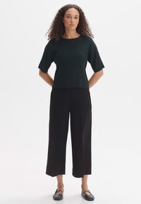 Un haut à manches courtes côtelé vert foncé associé à un pantalon large noir et des chaussures noires à enfiler, sur fond blanc.