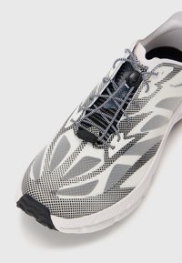 Chaussure de sport blanche avec une tige en mesh ornée d'un motif à pois noirs, système de laçage intégré et semelle texturée grise.