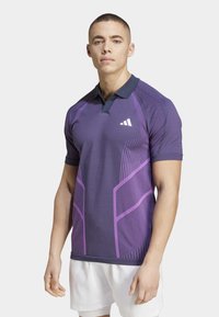 Polo shirt atlético morado con cuello navy, que presenta patrones geométricos en púrpura más claro, mangas cortas y un diseño ajustado.