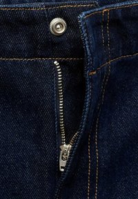 Tamno plava denim tkanina prikazuje krupni plan zatvarača s metalnim srebrnim povlačenjem i kontrastnim narančastim šavovima uz rubove.