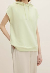 Sudadera sin mangas de color verde claro, hecha de tela suave, con un cordón en el cuello y un ajuste relajado. Combinada con pantalones beige.