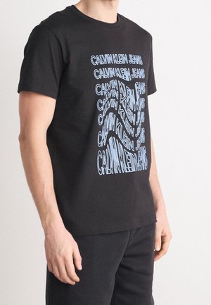 Mand iført sort t-shirt og bukser, t-shirten har gentaget forvrænget blå tekst "Calvin Klein Jeans" på forsiden, stående mod en hvid baggrund.