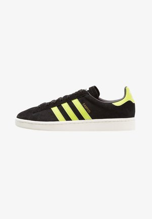 Zapatillas de ante negro con tres rayas reflectantes amarillas. Cuentan con una suela de goma blanca, cordones negros y un logo cerca del talón.