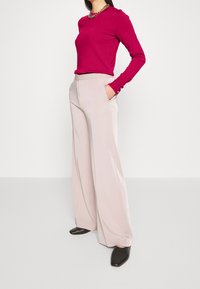 Fuchsia top met lange mouwen gecombineerd met lichtbeige broek met wijde pijpen, voorzien van zakken en een gladde textuur. Zwarte slip-on schoenen maken de look compleet.