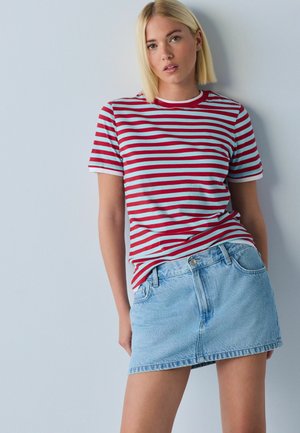 Blonde Frau lehnt an der Wand, trägt ein rot-weiß gestreiftes T-Shirt und einen hellblauen Jeans-Minirock, blickt mit neutralem Gesichtsausdruck nach vorne.