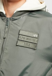 Vihreä pommikonesati, jossa on vetoketjukiinnitys, ribattu kaulus ja tikattu merkki, jossa lukee "Alpha Industries" ja "1959". Silkkinen pinta.