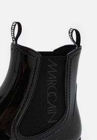 Bottine noire avec finition laquée brillante, panneaux élastiques sur les côtés et une languette de tirage, arborant le logo "Marc Cain" sur le textile.