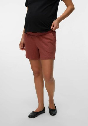 Person wearing rust-colored elastic waist shorts, black top, and black ballet flats, standing with one hand adjusting the shorts waistband.