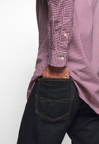 Košile s červenobílým kostkovaným vzorem a knoflíky, nošená přes tmavé džínové kalhoty s žlutým šitím na zadních kapsách.