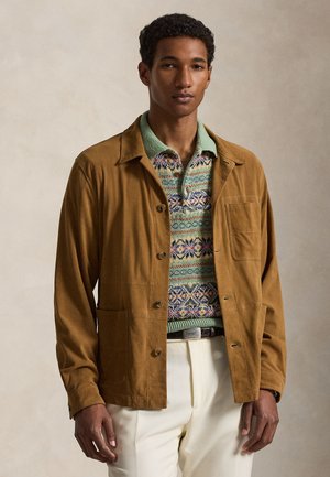 Młody mężczyzna ubrany w brązową zapinaną na guziki kurtkę, pod nią wzorzysty zielony sweter typu polo oraz jasno-kremowe spodnie, stojący na neutralnym tle.