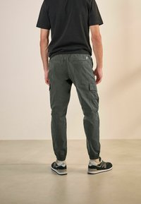 Tamsiai pilkos spalvos cargo kelnės su elastiniais manžetais, šoninėmis kišenėmis ir siuvinėtų detalių, vilkimos juodais New Balance sportbačiais.