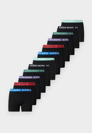 Schlanker Mann in schwarzer Björn Borg Unterwäsche steht barfuß auf einem Teppich und legt die Hand auf einen Stuhl mit Chromrahmen und grauem Sitz und Rückenlehne.