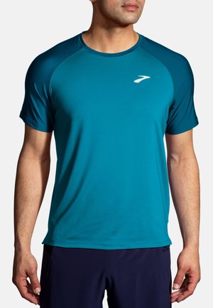 Homme portant un t-shirt de sport à manches courtes couleur sarcelle avec un petit logo blanc sur la poitrine gauche et un short foncé.
