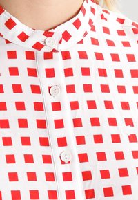 Valkoinen paita punaisilla neliökuvioilla, nappilista edessä ja pieni brodeerattu logo oikealla puolella lähellä rintaa.
