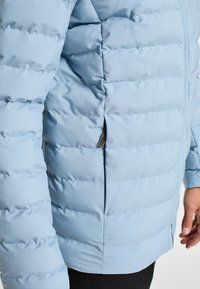 Lysblå dunjakke med quiltet design, der har en lynlåslommer i siden og en glat tekstur. Nærbillede, der fremhæver stof og form.