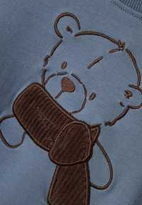 Tessuto azzurro chiaro con un orso ricamato marrone che tiene una sciarpa di corduroy marrone, con elementi lisci e testurizzati nel design.
