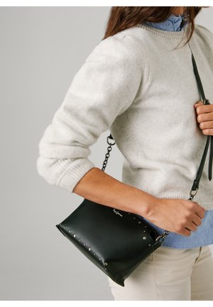 Vrouw draagt witte trui en blauw overhemd en draagt een zwarte leren crossbodytas met metalen studs en kettingriem.