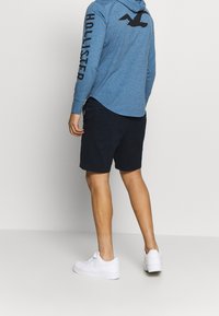 Man med blå långärmad hoodie med "Hollister"-text på ärmen, svarta shorts och vita Nike-sneakers som står mot en enfärgad bakgrund.