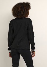 Musta bleiseri, jossa istuva malli, näkyvillä oleva sauma ja kontrastiset raidalliset kalvosimet. Sileä kangas ja jäsennelty siluetti.