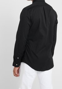 Černá košile s dlouhými rukávy na knoflíky, s oblouhovým lemem, bočními rozparky a záhybem na zádech. Vyrobeno z lehké látky.