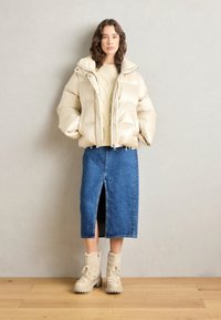 Beżowa kurtka puchowa, kremowy sweter z dzianiny, dżinsowa spódnica midi z przednim rozcięciem oraz beżowe botki z ozdobami z kożucha. Gładkie kolory, casualowy styl.