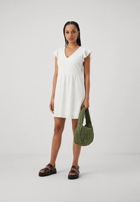 Bela bombažna obleka s kratkimi volanastimi rokavi, izrezom v obliki črke V, detajli z luknjicami, razširjenim krilom, kombinirana z zeleno prešito torbico in črnimi sandali.