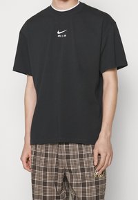 Svart bomulls-T-shirt med rund hals, korta ärmar och vitt Nike AIR-logotyp på bröstet. Kombinerad med bruna rutiga byxor.