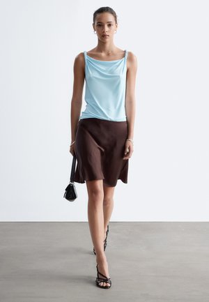 Femme marchant portant un haut sans manches bleu clair, une jupe marron foncé, des talons noirs à brides et portant un petit sac à main noir sur un fond uni.