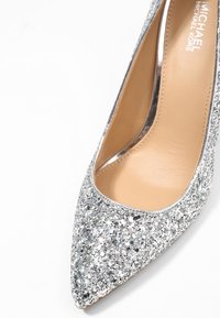 Chaussure à talon haut pailletée argentée avec un design à bout pointu, dotée d'une semelle intérieure beige lisse et d'une bordure métallique le long des bords.