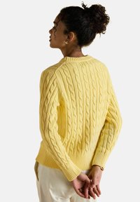 Moteris su garbanotais plaukais surišta į kuodą, vilkinti geltoną džemperį su raštu ir baltas kelnes, stovinti su įremtomis rankomis už nugaros.