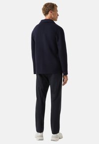 Granatowa kurtka wełniana z domieszką, o wyraźnym kołnierzu, eleganckim fasonie i guzikami przy mankietach, w zestawieniu z ciemnymi spodniami i jasnymi trampkami.