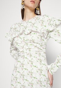 Vestido floral blanco con mangas acampanadas y volantes, que presenta patrones en rosa y verde. Tejido texturizado con detalles fruncidos a lo largo de la cintura.