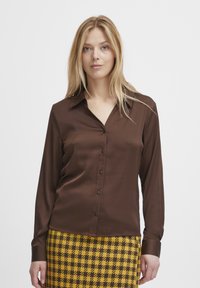 Braune Satinbluse mit langen Ärmeln und einem leicht offenen Kragen, ausgestattet mit einer durchgehenden Knopfleiste. Dazu ein Rock im Hahnentrittmuster in Gelb und Schwarz.