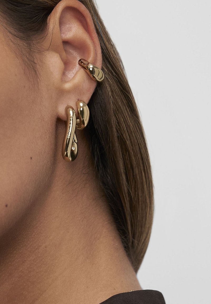 Nærbillede af et højre øre, der bærer to guldøreringe i bølget form, en på øreflippen og en på den øverste bruskdel af øret.