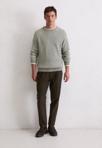 Pull en tricot gris clair à col rond et manches côtelées, associé à un pantalon olive foncé et des chaussures marron. Se tenant devant un fond uni.