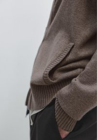 Brauner Pullover aus weichem Material mit gerippten Bündchen an Ärmeln und Saum. Seitliche Taschen sind sichtbar, getragen über schwarzer Hose.