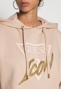 Felpa rosa chiaro con logo triangolare e testo "GUESS". Presenta dettagli dorati sui lacci e un ricamo testurizzato.