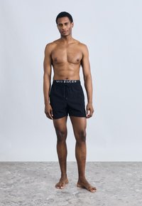 Μαύρα μαγιό σορτς με κορδόνι στη μέση, με ελαστική ζώνη μέσης με λογότυπο. Το μοντέλο στέκεται ξυπόλυτο μπροστά σε απλό φόντο.