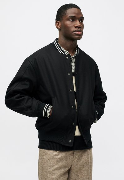 Νεαρός άνδρας με μαύρο μπουφάν μπάσκετ πάνω από πουλόβερ με ρίγες και καρό πουκάμισο, με μπεζ υφασμάτινο παντελόνι, χέρια στις τσέπες.