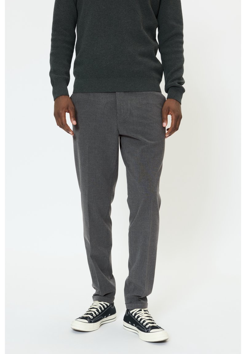 Homme portant un pull gris foncé, un pantalon de costume gris et des baskets basses noires avec des lacets blancs, debout devant un fond blanc.