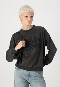 Felpa grigia con la parola "CRUSH" in lettere nere in rilievo, colletto e polsini a costine, abbinata a jeans in denim blu chiaro e a una borsa nera.