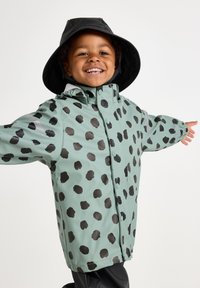 Grøn vandtæt regnfrakke med store sorte prikker, der har trykknaplukning og lange ærmer. Båret med en sort bredskygget hat.