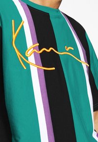 Zielona, czarna i fioletowa koszulka w paski z pomarańczowym haftowanym logo. Krótkie rękawy i okrągły dekolt. Wykonana z miękkiego materiału.