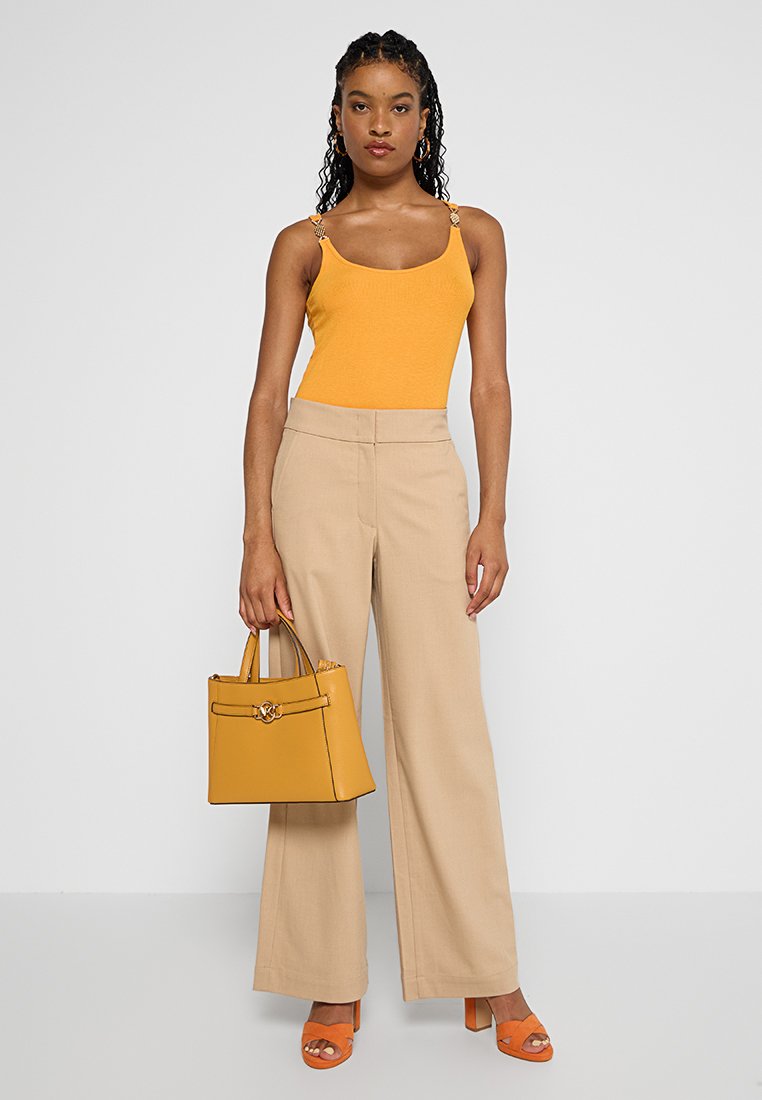 Femme portant un débardeur moutarde, un pantalon beige à jambes larges, des talons oranges, portant un sac à main moutarde sur fond blanc uni.