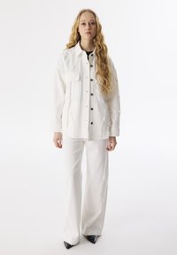 Giacca bianca con cuciture visibili e grande tasca frontale, abbinata a pantaloni bianchi coordinati e tacchi a punta neri.
