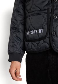 Veste matelassée noire avec un motif texturé, dotée d'une poche avant et d'un détail brodé "RR.2013:01". Les poignets côtelés complètent le design.