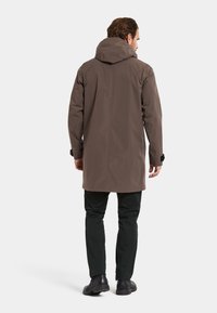 Brun vandtæt jakke med hætte, lange ærmer, justerbare manchetter og lige kant. Blevet parret med mørke bukser og sorte støvler.